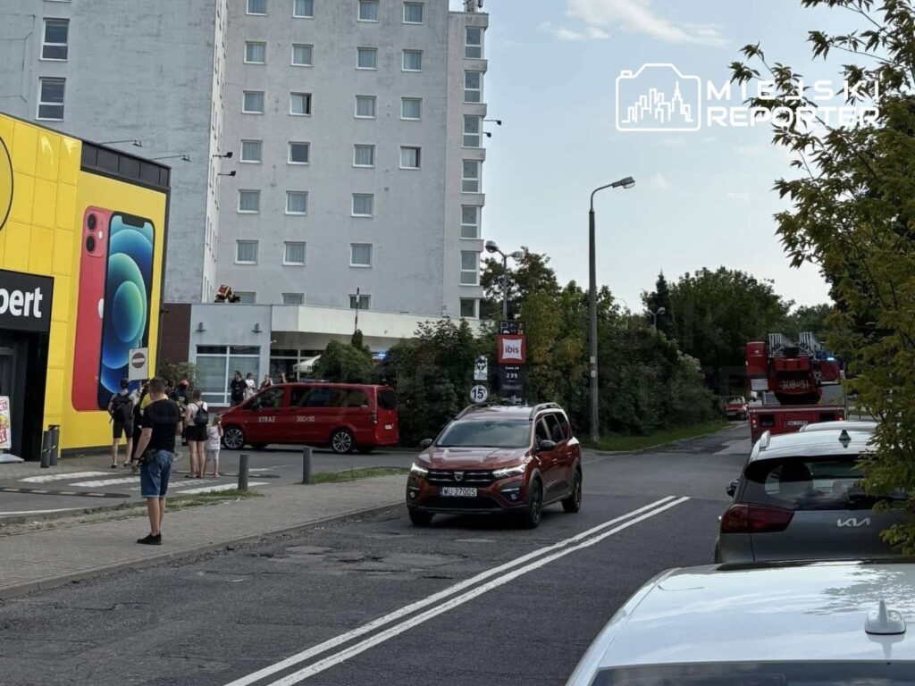 Grupa osób obserwuje interwencję służb ratunkowych przed nowoczesnym hotelem, w tle widoczny czerwony wóz strażacki.