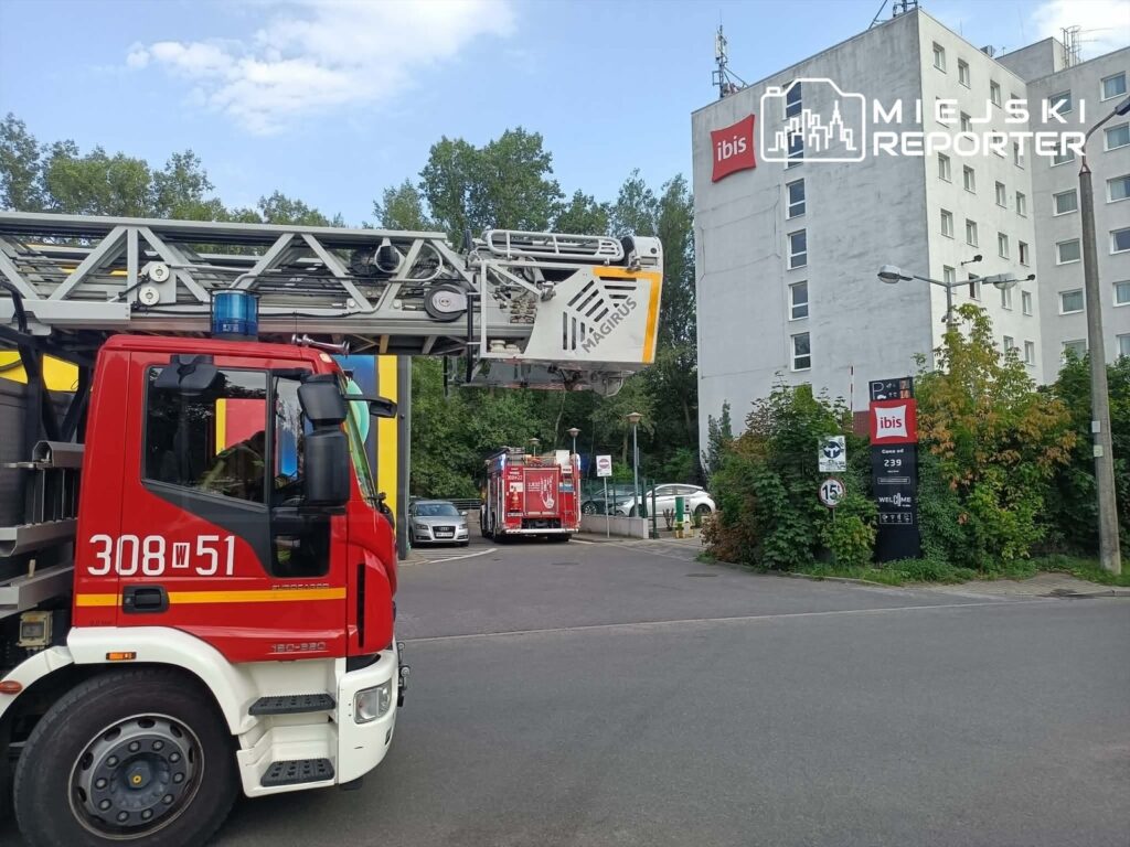 Czerwony wóz strażacki z podnośnikiem stoi przed nowoczesnym hotelem, obok widoczny drugi wóz strażacki.