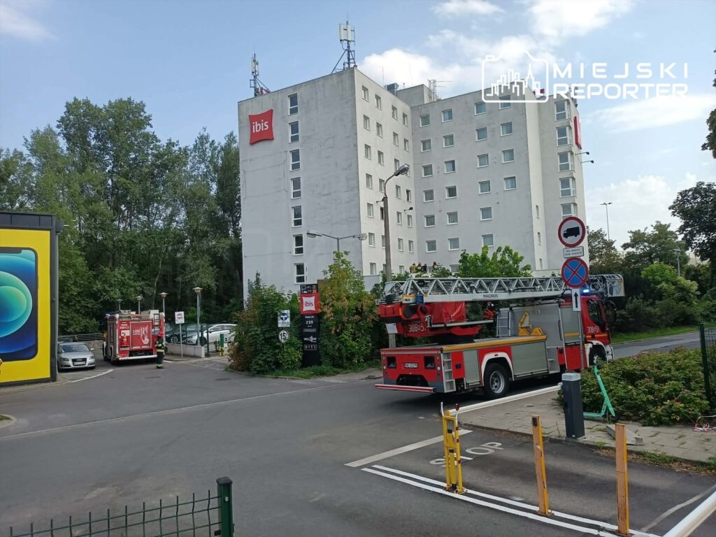 W tle nowoczesny hotel z logo, a na pierwszym planie wóz strażacki z drabiną, zaparkowany obok zieleni.