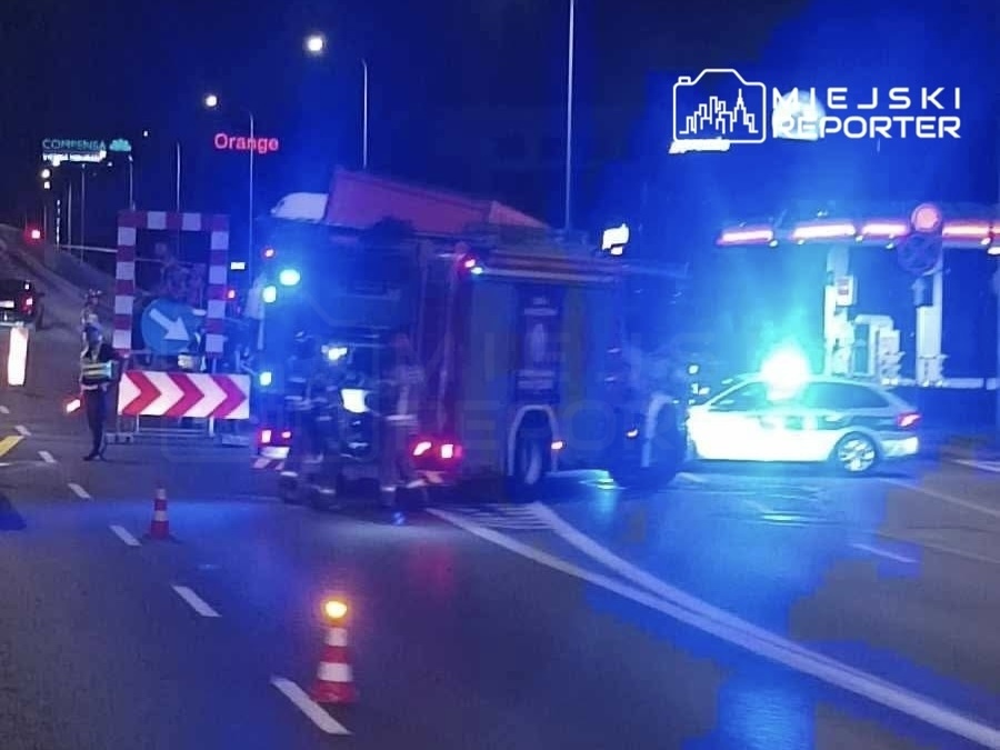 W nocy na drodze stoi woz strażacki z włączonymi sygnałami świetlnymi, obok pracują policjanci kierujący ruchem.