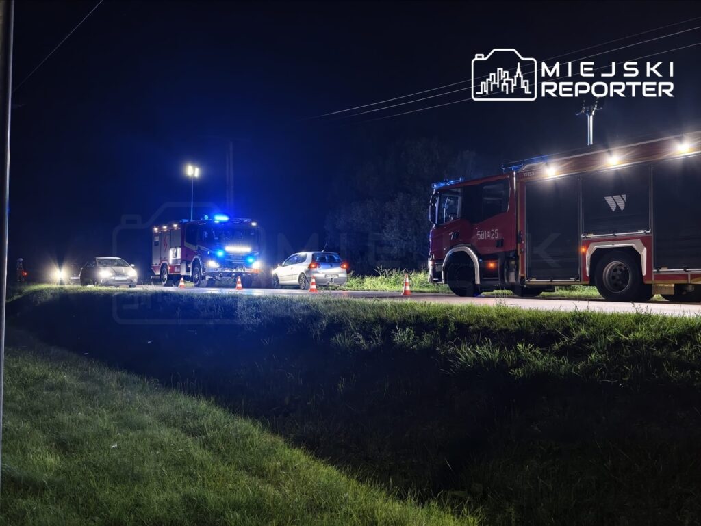Na drodze nocą, straż pożarna z włączonymi sygnałami świetlnymi zabezpiecza miejsce zdarzenia z udziałem kilku pojazdów.