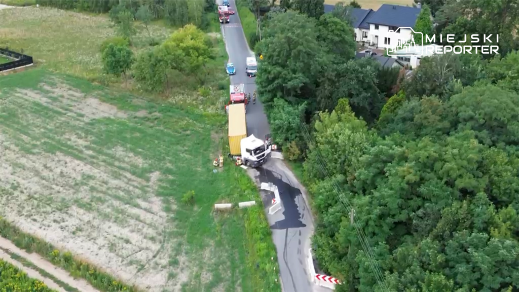 Ciężarówka z żółtym kontenerem zablokowała drogę, a obok stoją pojazdy straży pożarnej i ambulans. W tle zieleń i domy.