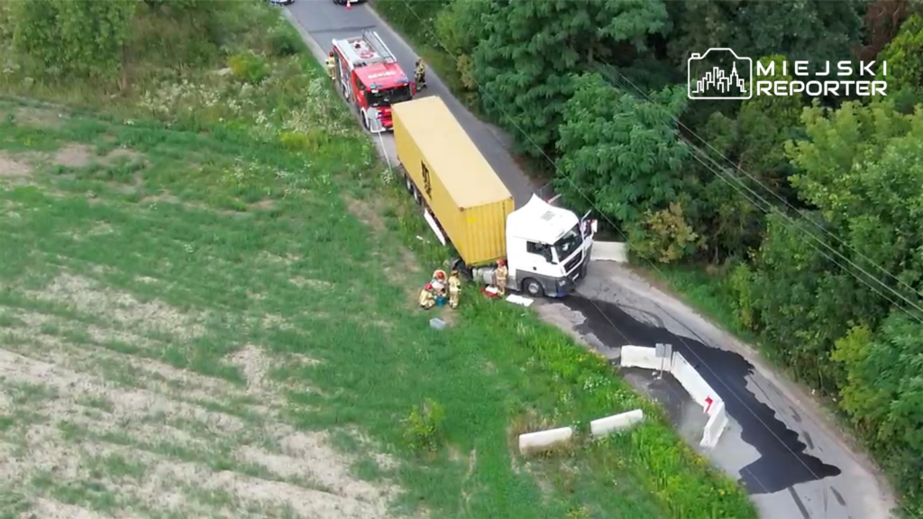 Ciężarówka z żółtym kontenerem stoi przy drodze, obok której pracują strażacy, a na jezdni widoczne są betonowe bariery.