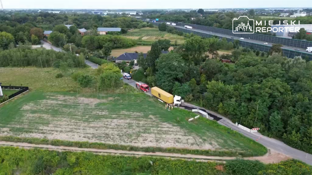 Ciężarówka z żółtą naczepą stoi przy zablokowanej drodze, obok interweniują strażacy i policja. W tle zieleń i zabudowania.