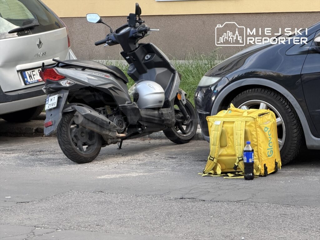 Skuter zaparkowany obok samochodów, obok niego żółta torba dostawcza Glovo z butelką napoju na chodniku.