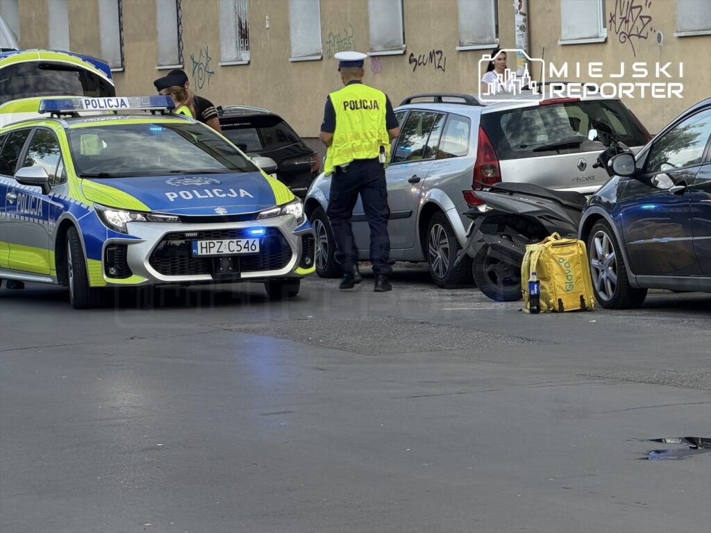 Policjanci w odblaskowych kamizelkach badają kolizję drogową w miejskiej okolicy, obok uszkodzonych samochodów.