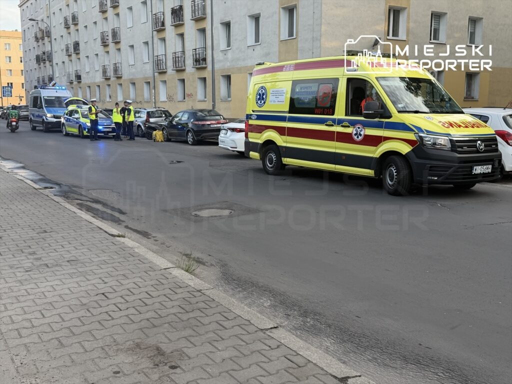 Grupa policjantów w odblaskowych kamizelkach rozmawia obok ambulansu i radiowozu na ulicy w mieście.