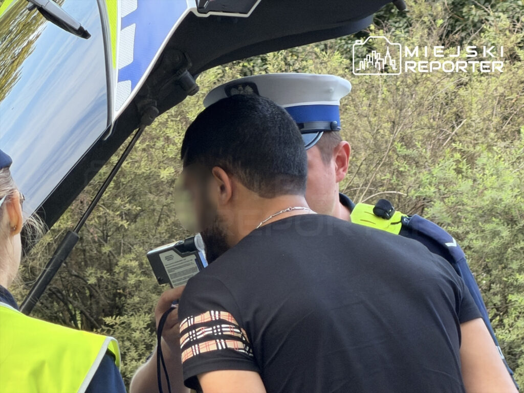 Mężczyzna w czarnej koszulce rozmawia z policjantem przy otwartym bagażniku radiowozu.
