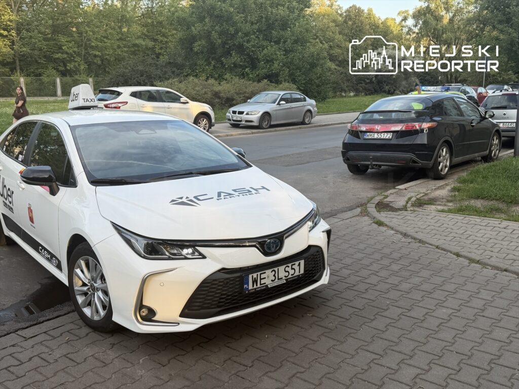 Białe taksówki z logo Ubera i Cash czekają na pasażerów na parkingu obok jezdni, w tle widać inne samochody.