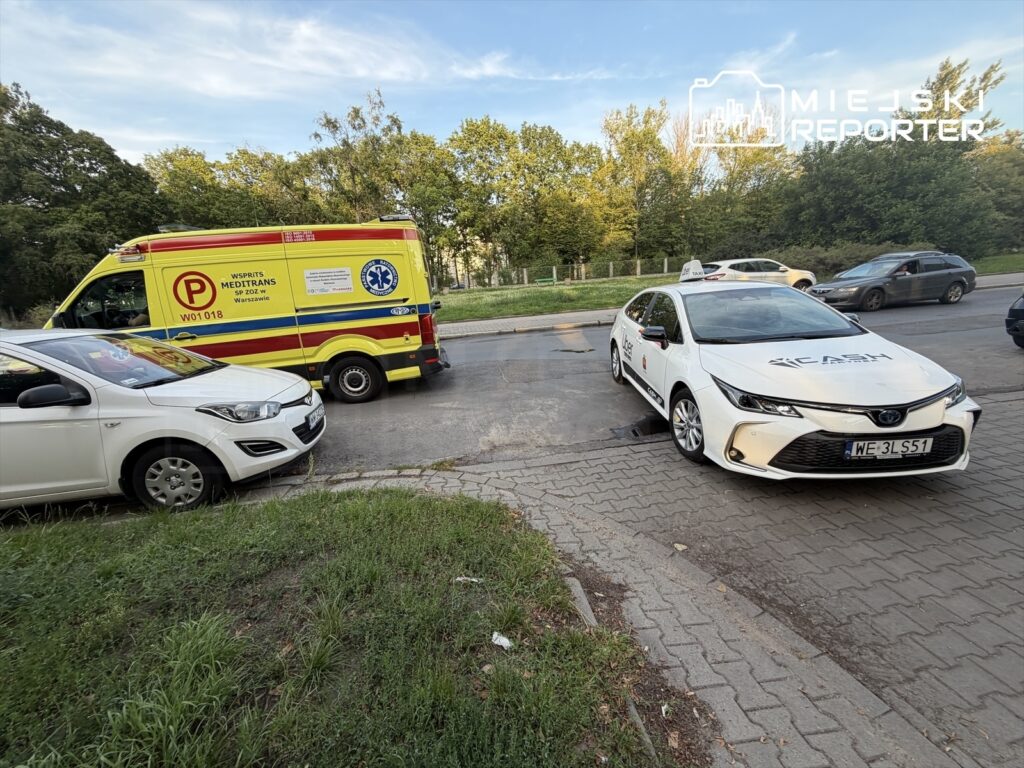 Ambulans z charakterystycznymi oznaczeniami zatrzymany na ulicy obok białego samochodu osobowego i taksówki.
