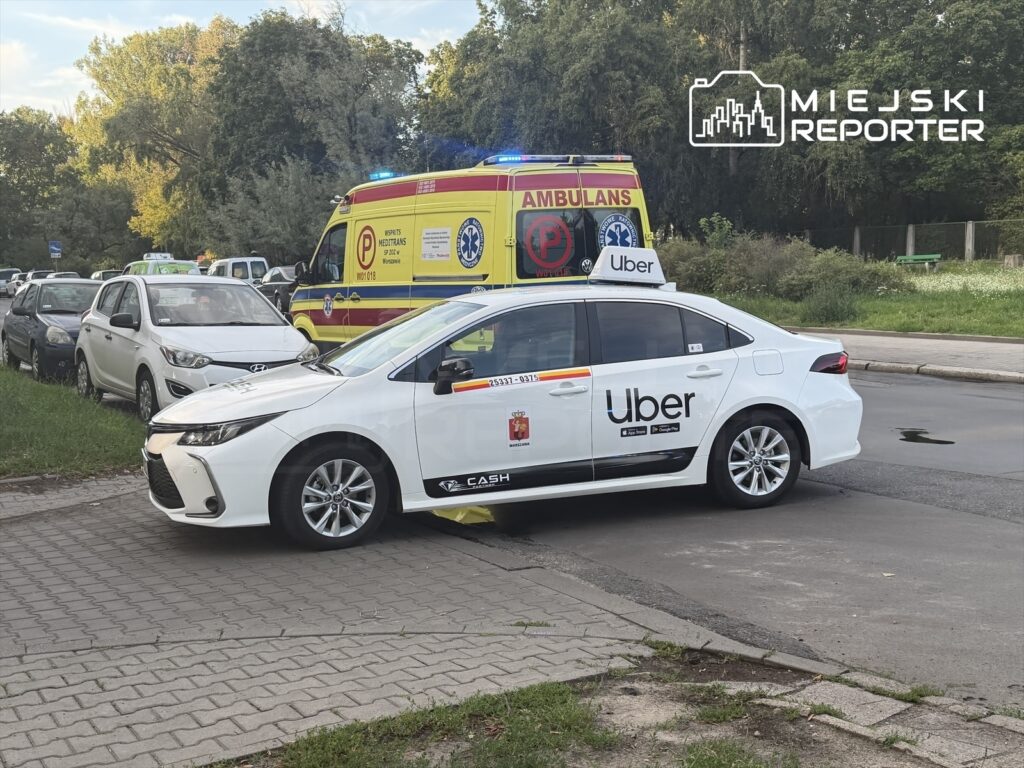 Samochód osobowy z logo Ubera stoi na chodniku obok karetki pogotowia z włączonymi sygnałami świetlnymi.