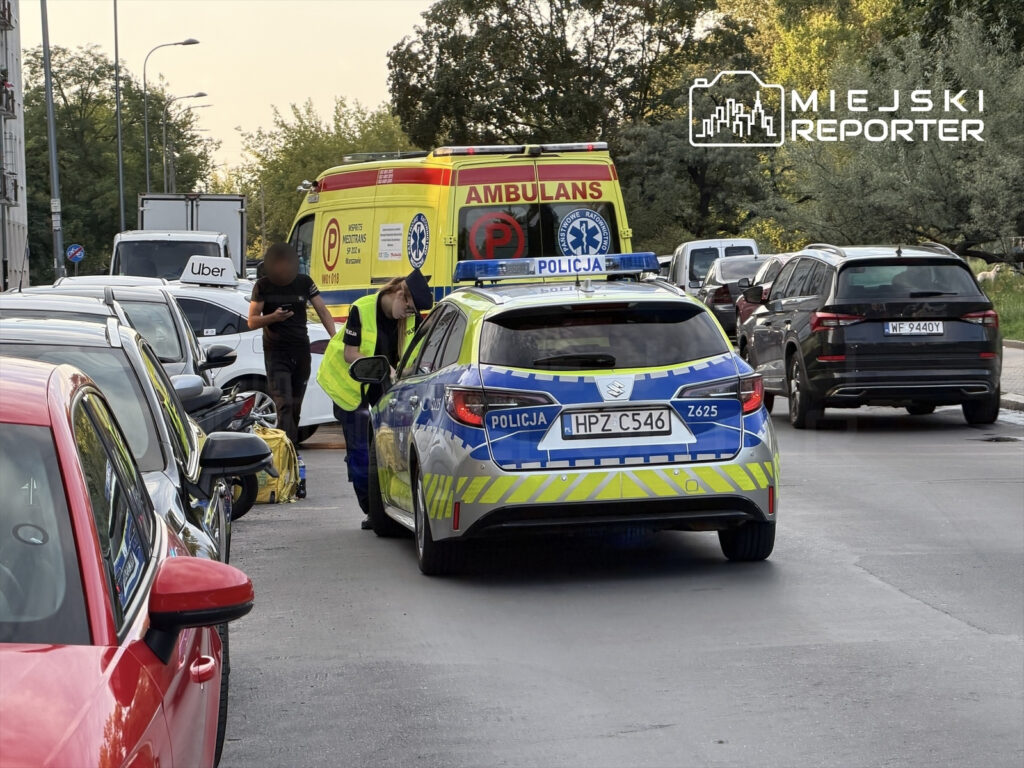 Policjant w neonowej kamizelce sprawdza dokumenty w radiowozie, obok karetka pogotowia oraz zaparkowane samochody.