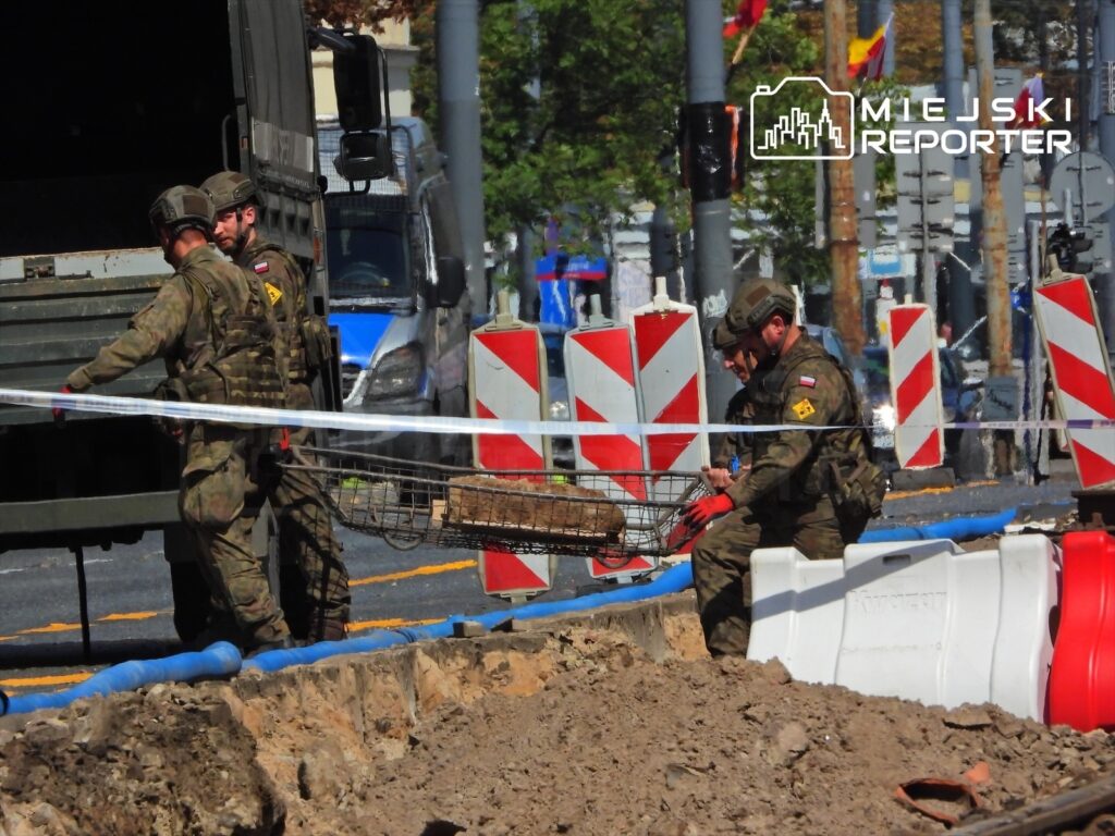 Czterech żołnierzy w mundurach wojskowych pracuje przy wykopie, przenosząc przedmioty w koszu. W tle widoczne znaki drogowe i pojazdy.