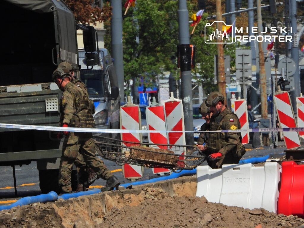 Dwaj żołnierze w mundurach wojskowych przenoszą metalowy kosz na placu budowy, otoczonym barierkami.