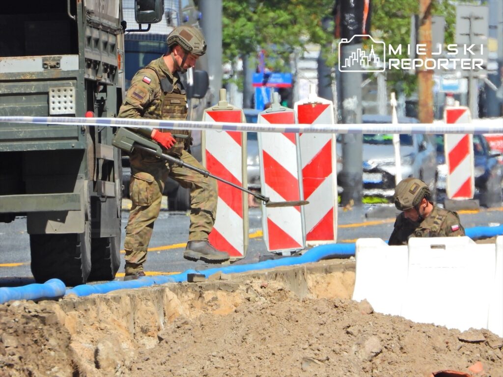 Żołnierz w mundurze z bronią w ręku patroluje teren budowy, podczas gdy drugi żołnierz pracuje w wykopie.