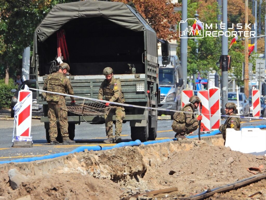 Żołnierze w mundurach rozładują sprzęt z wojskowego pojazdu w rejonie wykopu na ulicy.