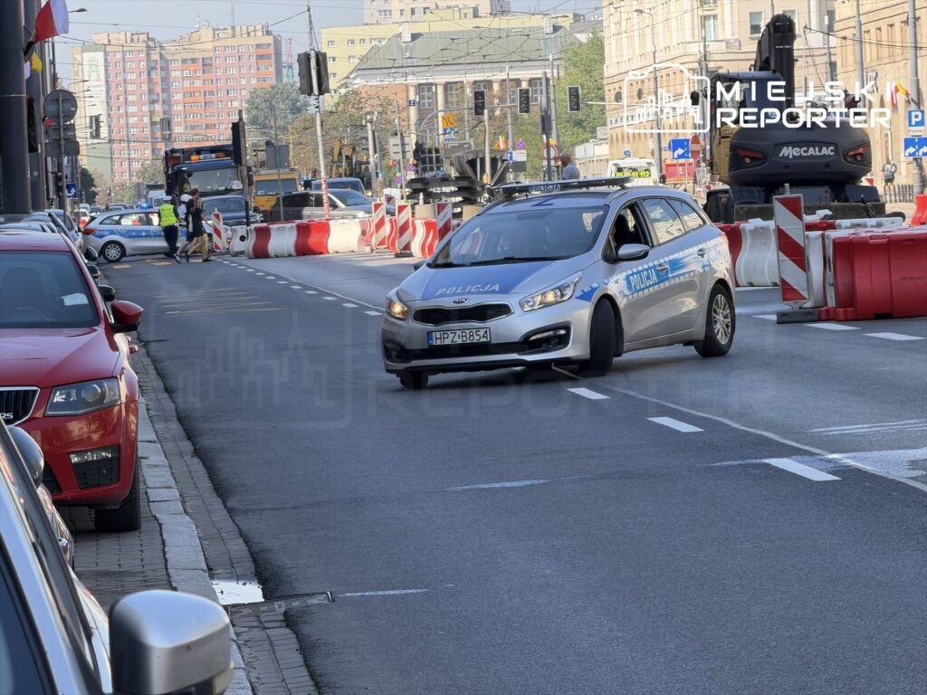 Policyjny radiowóz przejeżdża obok zamkniętej alei, a w tle widać pojazdy budowlane i sygnalizację świetlną.