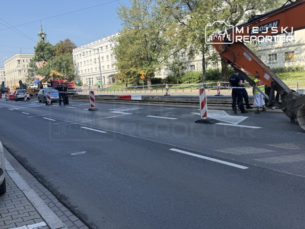 Policjanci rozmawiają z pracownikami przy maszynie budowlanej na zamkniętej ulicy w centrum miasta.