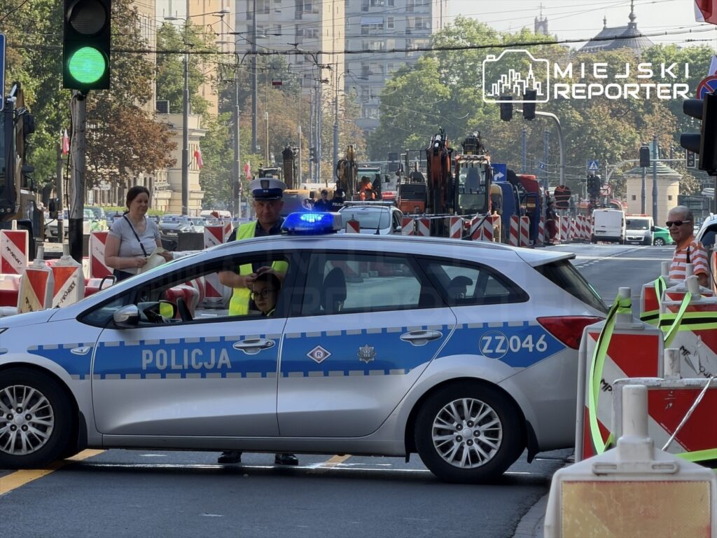 Policjant w kamizelce odblaskowej kieruje ruchem obok radiowozu, na tle zamkniętej drogi z barierkami.