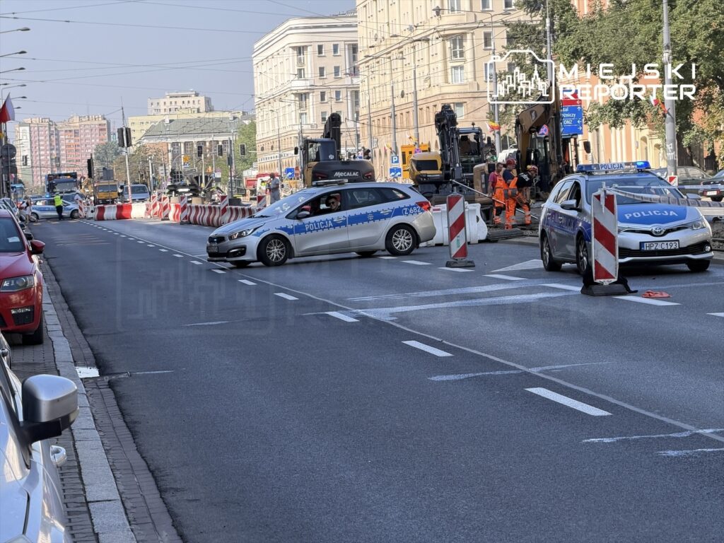 Policjanci kierują ruchem drogowym na zamkniętej Aleji „Solidarności”, obok pracowników budowlanych.