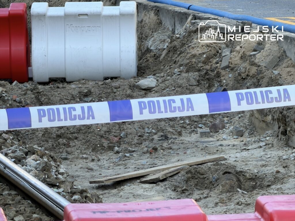 Wykop w ziemi z oznaczeniem policji, w tle widoczne czerwone i białe pojemniki oraz leżące fragmenty materiałów budowlanych.