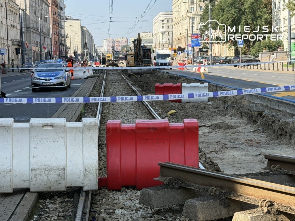 Policjant w oznakowanym radiowozie monitoruje prace budowlane przy zamkniętej alejce, otoczonej barierkami i taśmą policyjną.