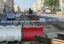 Policjant w oznakowanym radiowozie monitoruje prace budowlane przy zamkniętej alejce, otoczonej barierkami i taśmą policyjną.