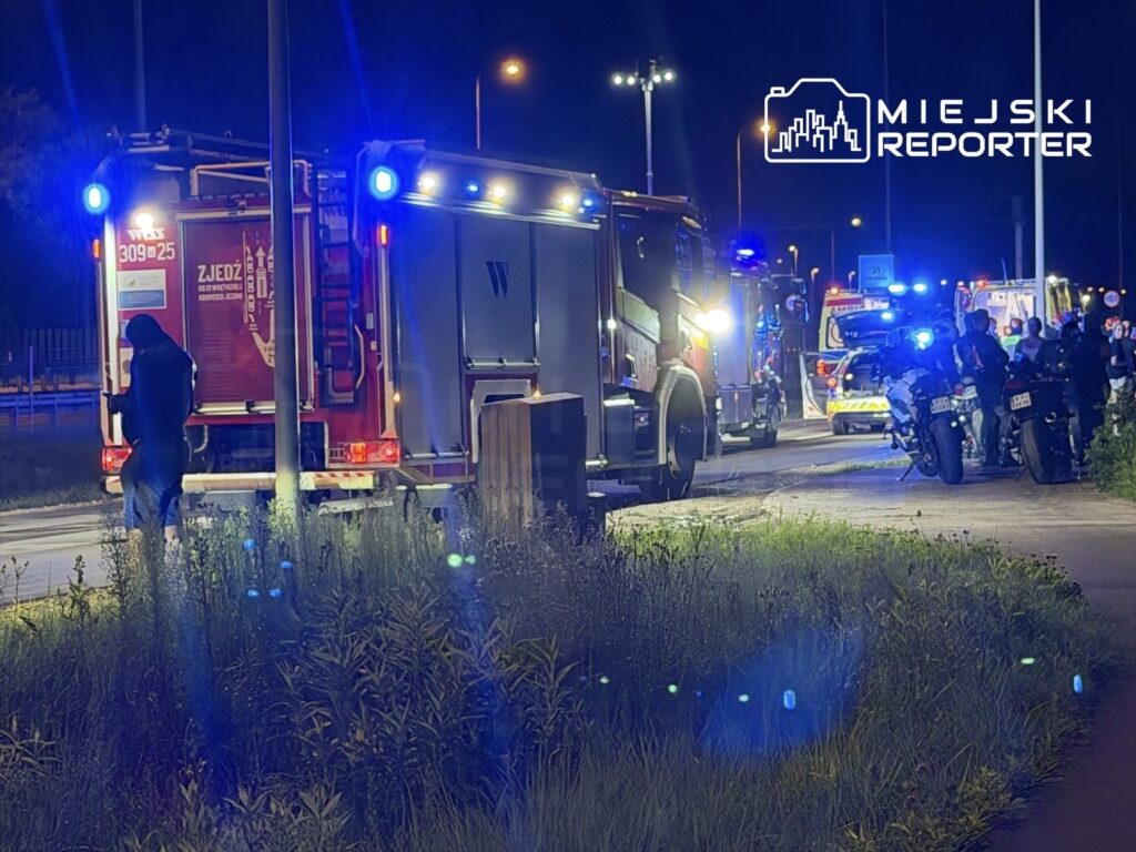 Na drodze zaparkowane są pojazdy służb ratunkowych z włączonymi sygnałami świetlnymi, a w tle widać grupę osób.