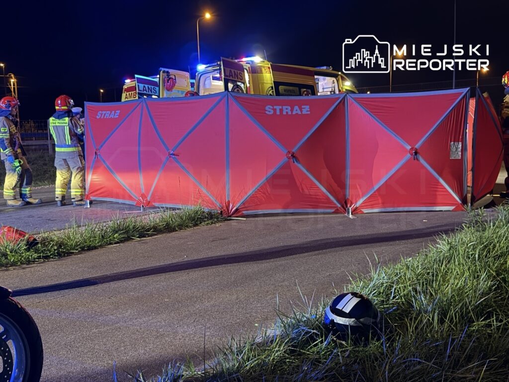 Grupa strażaków w mundurach pracuje przy rozłożonym czerwonym namiocie na poboczu drogi w nocy, obok pojazdów ratunkowych.