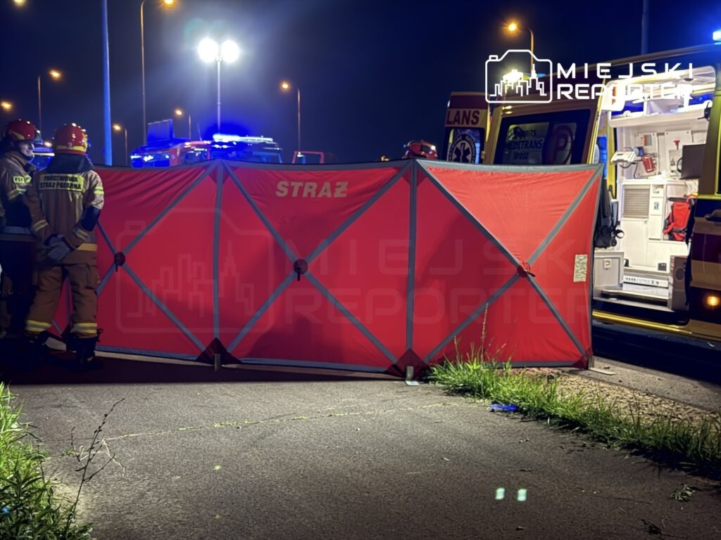 Grupa strażaków w mundurach stoi przed czerwonym namiotem medycznym na oświetlonej ulicy, obok karetki.