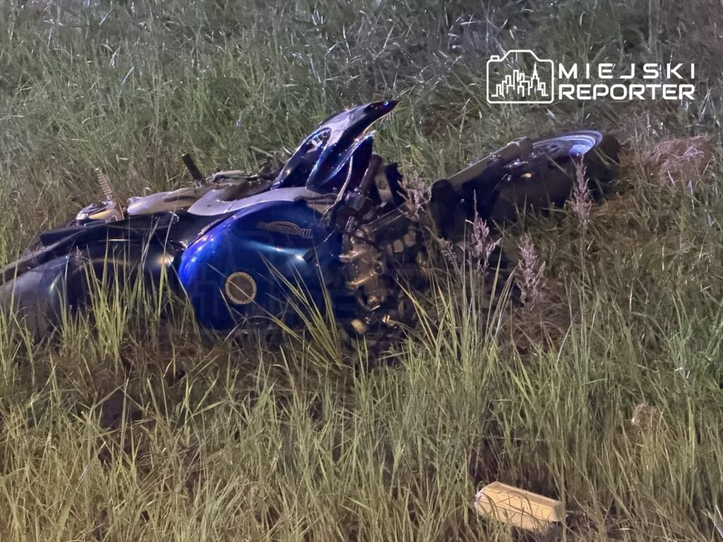 Zniszczony motocykl leży na trawie przy drodze, otoczony wysoką roślinnością.