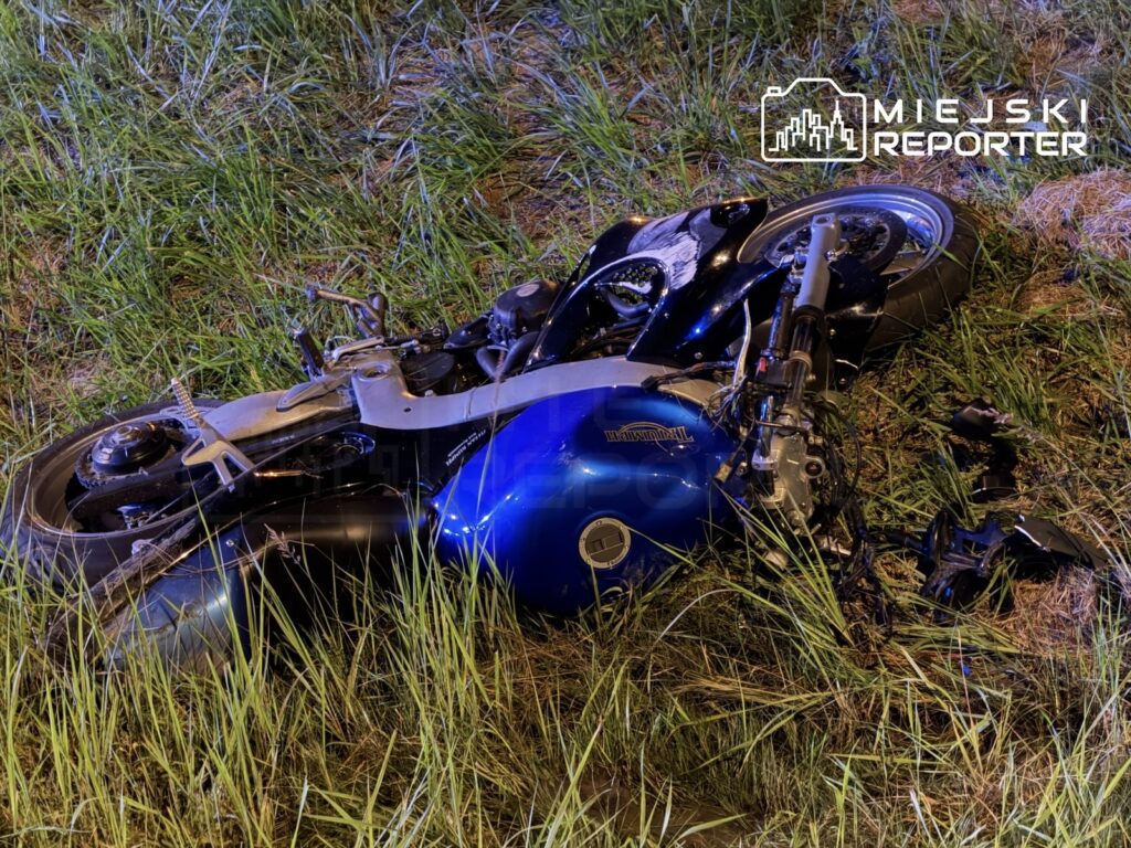 Motocykl leżący na trawie, z uszkodzoną karoserią i odłączonymi elementami, w nocy.