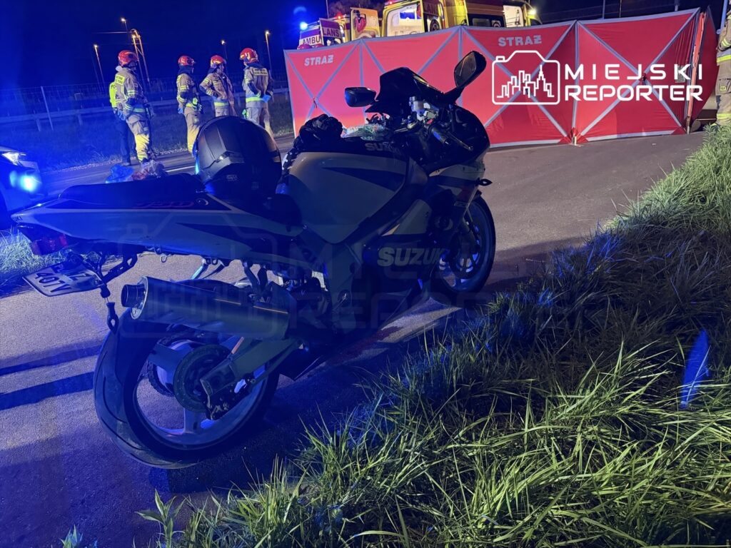 Motocykl marki Suzuki stoi na poboczu drogi, w tle widoczni strażacy przy czerwonym namiocie.