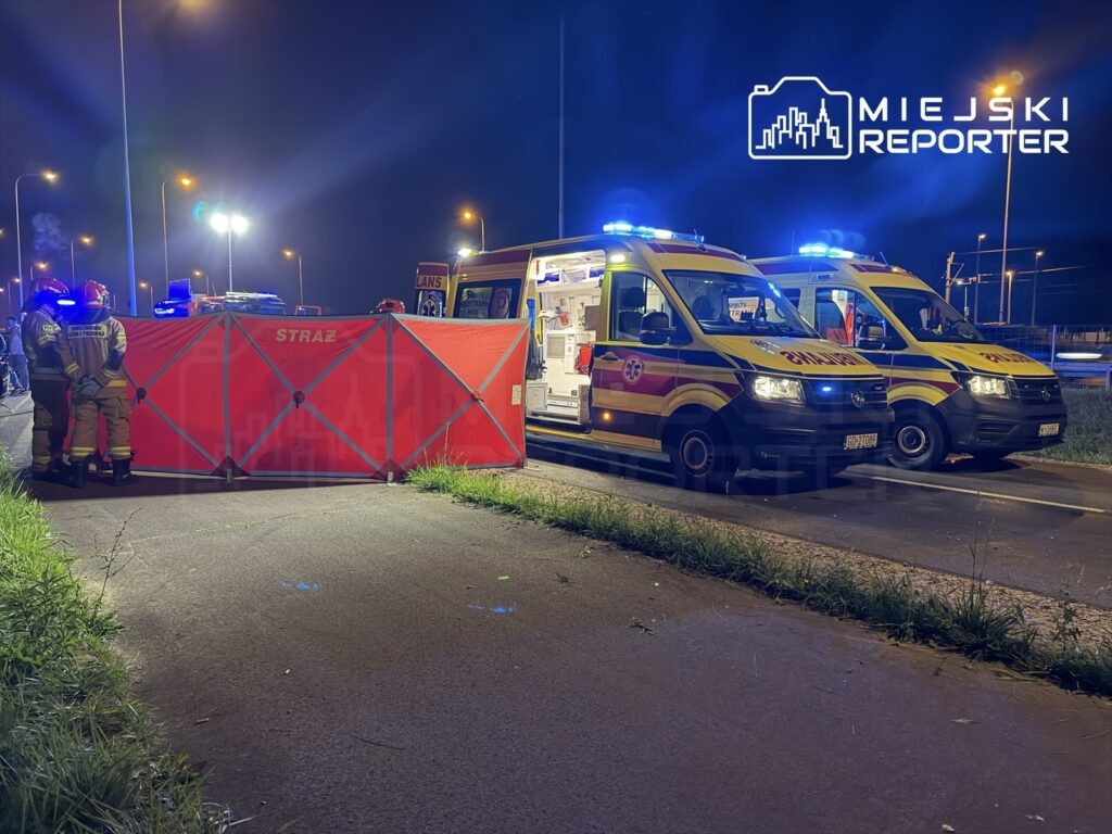 Ratownicy medyczni stoją przy czerwonej bariery zabezpieczającej, obok ambulansów z włączonymi sygnałami świetlnymi.