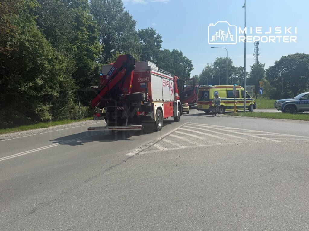 Na drodze stoi wóz strażacki z podnośnikiem, obok ambulans oraz radiowóz policji, w tle zieleń drzew.