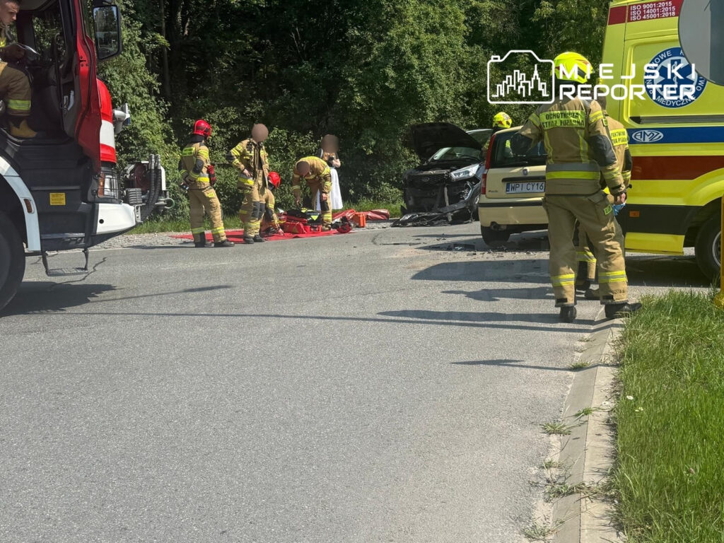 Ratownicy medyczni udzielają pomocy poszkodowanej 79-latce na drodze, obok zniszczonych pojazdów i karetki.