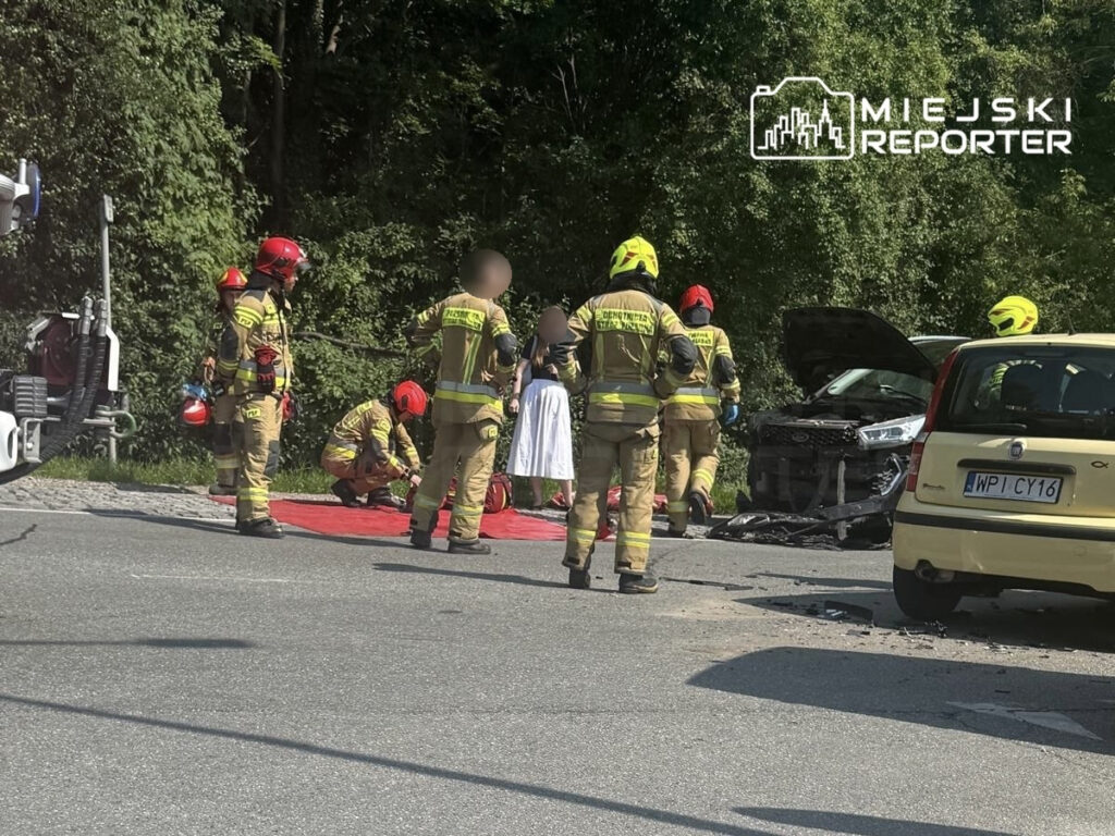 Grupa strażaków udziela pomocy poszkodowanej 79-latce na drodze, obok uszkodzonego samochodu.