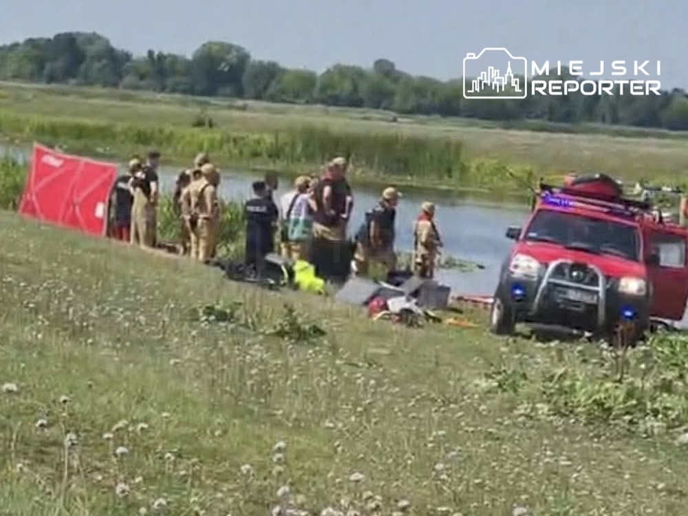 Grupa ratowników i strażaków na brzegu rzeki, obok czerwonego samochodu ratunkowego, przygotowuje się do akcji.