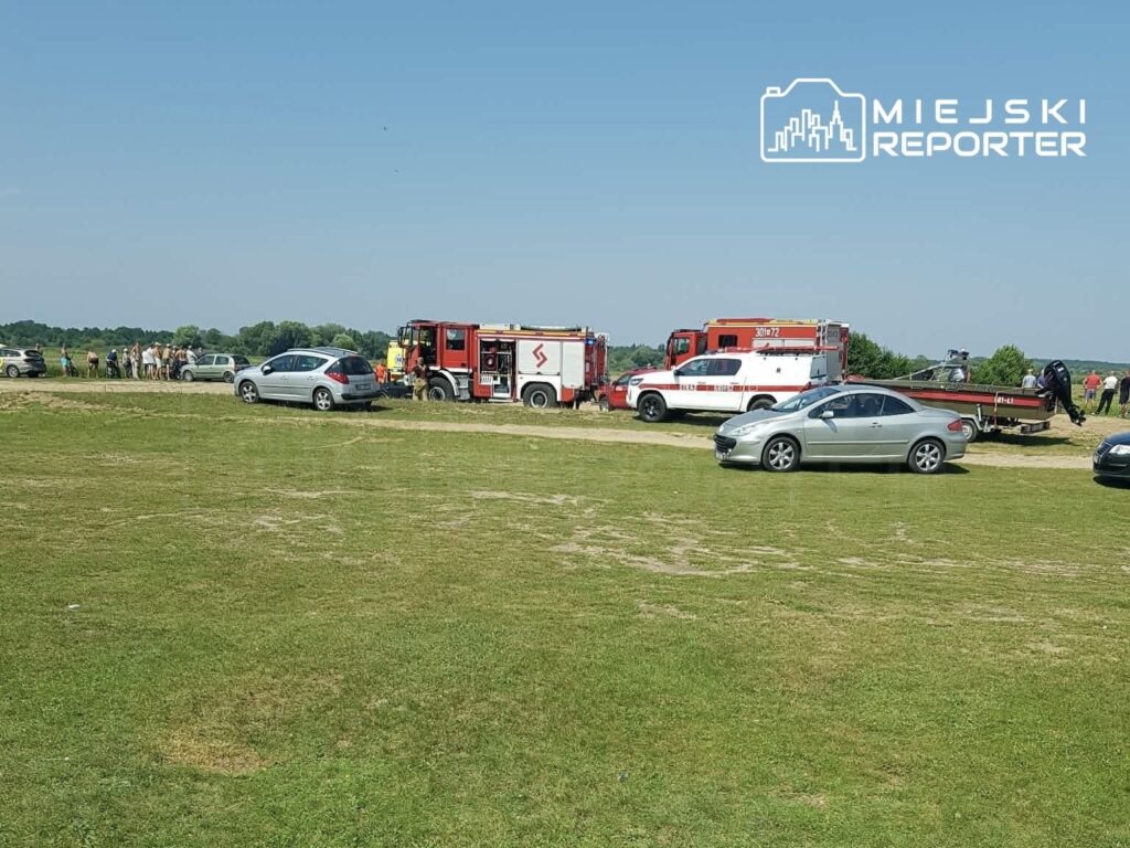 Grupa osób obserwuje działania służb ratunkowych przy wodzie, w tle widoczne wozy strażackie i ambulansy.