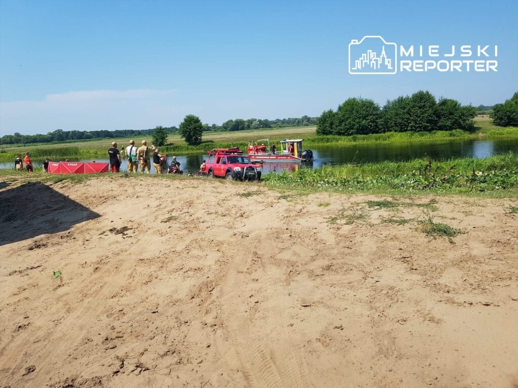 Grupa strażaków przygotowuje sprzęt ratunkowy nad brzegiem rzeki, w tle widać zieloną roślinność i spokojną wodę.
