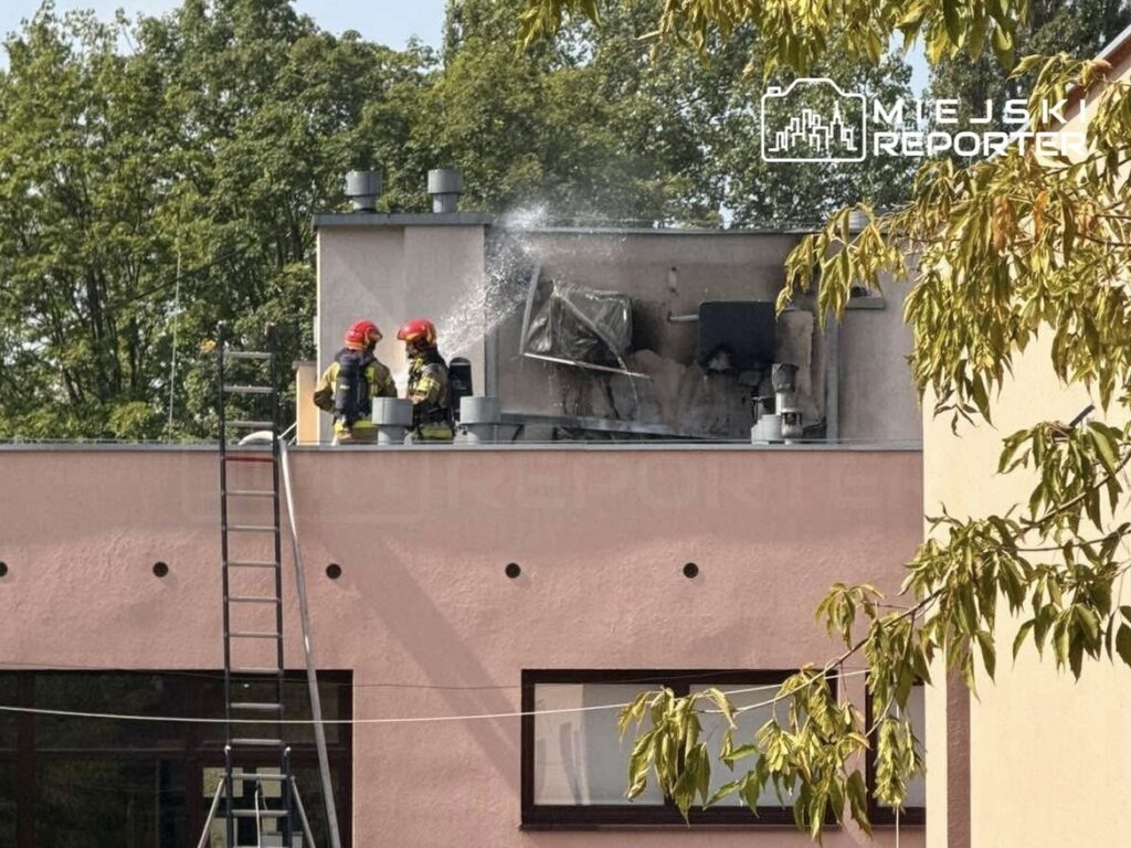 Dwóch strażaków w hełmach gasi pożar klimatyzatorów na dachu budynku, obok drabina. W tle drzewa.