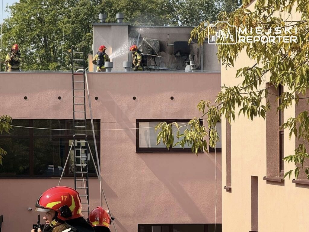 Strażacy w hełmach gaszą pożar klimatyzatorów na dachu budynku szkoły, używając węża strażackiego.