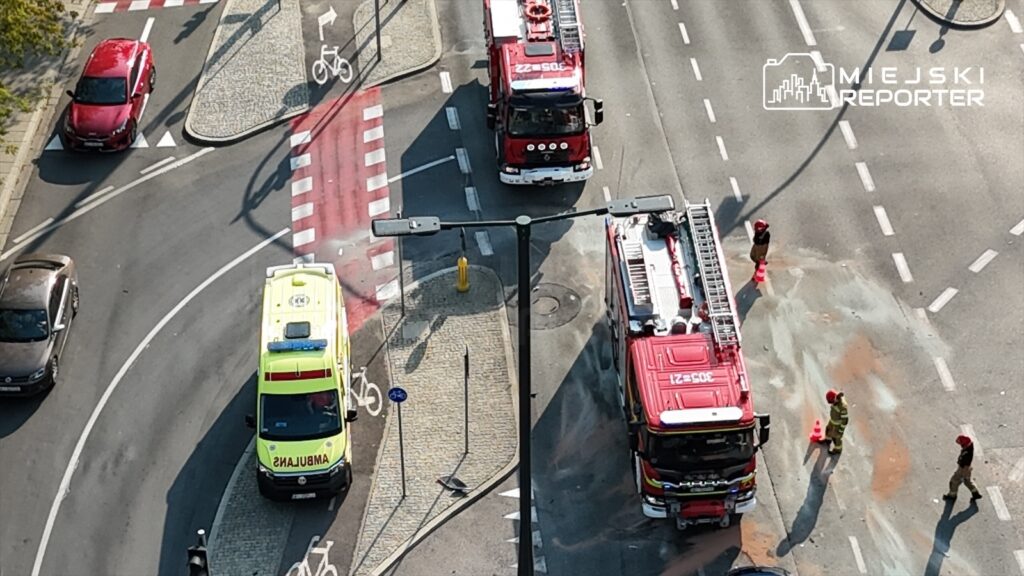 Ambulans i wóz strażacki na skrzyżowaniu, strażacy zabezpieczają miejsce zdarzenia, na jezdni widoczne plamy substancji.
