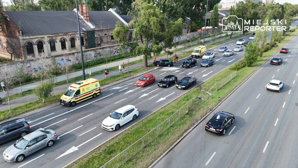 Karetka pogotowia zatrzymana na pasie ruchu, obok niej stoją samochody, a w tle widoczne budynki z graffiti.
