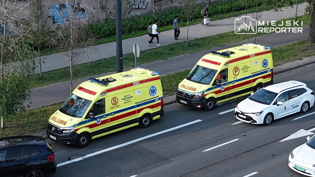 Dwa ambulanse z włączonymi sygnałami świetlnymi zaparkowane na poboczu drogi obok chodnika, obok przechodzący piesi.