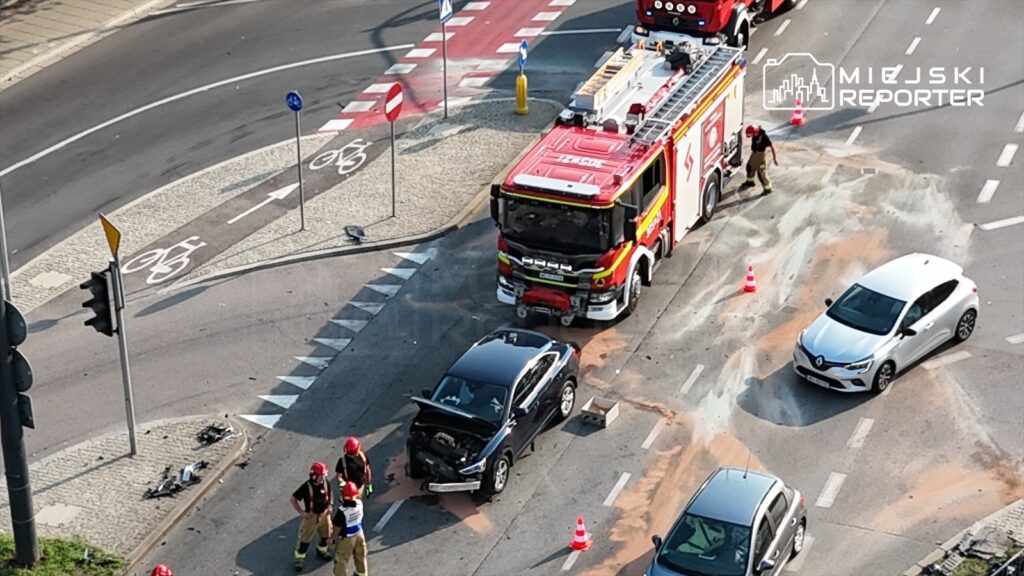 Czterech strażaków zabezpiecza miejsce wypadku, gdzie czarny samochód osobowy z uszkodzoną maską stoi na drodze, a obok znajduje się wóz strażacki.