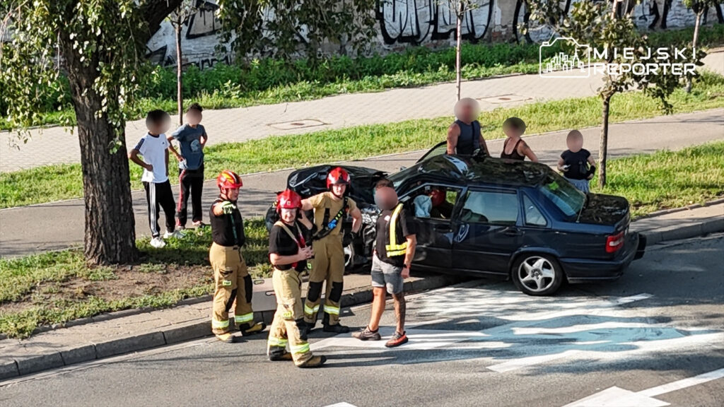 Grupa strażaków w kaskach rozmawia z osobami stojącymi obok uszkodzonego samochodu na drodze.