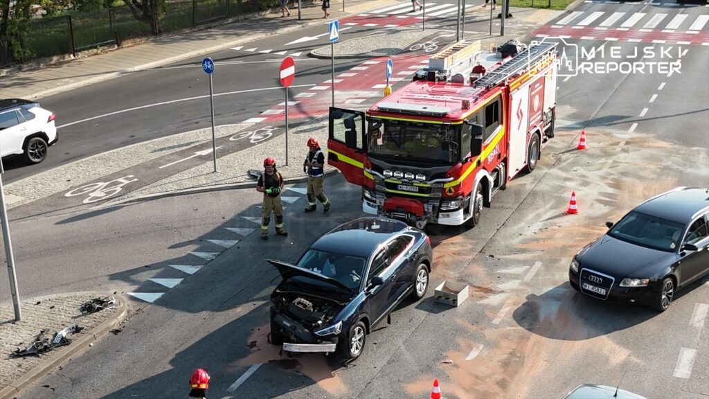 Czarny samochód osobowy z otwartą maską stoi na drodze, obok niego strażacy w hełmach rozmawiają z kierowcą.