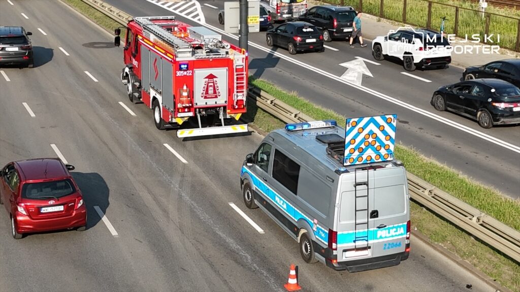 Czerwony wóz strażacki oraz radiowóz policyjny zatrzymane na drodze, obok zablokowane pojazdy osobowe.