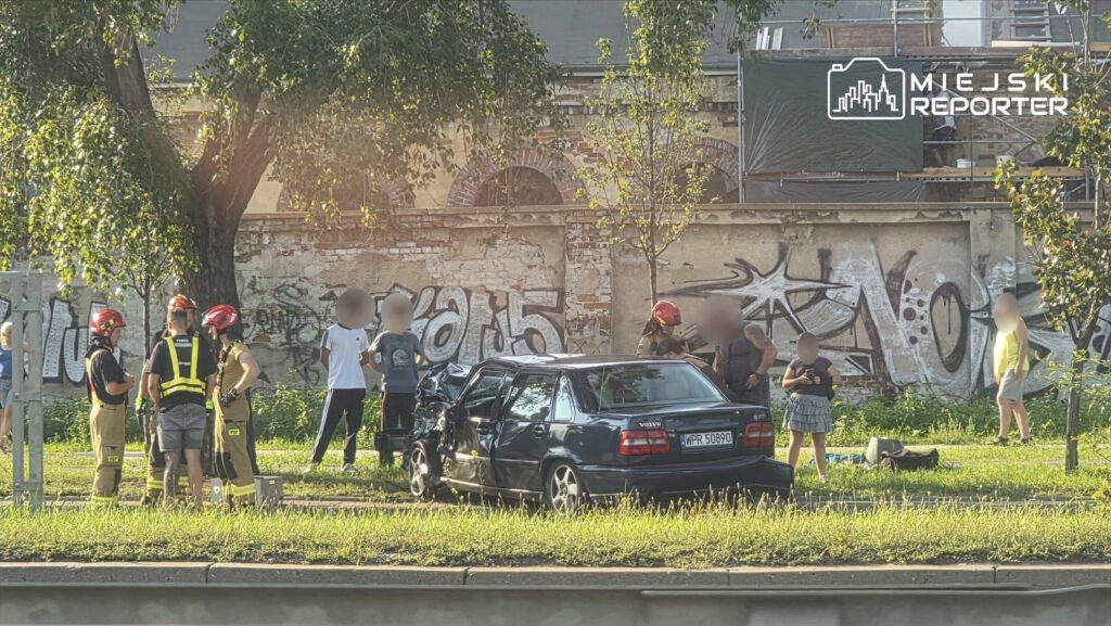 Na miejscu wypadku grupka ludzi obserwuje uszkodzone Volvo, obok pracują strażacy w kaskach i mundurach.